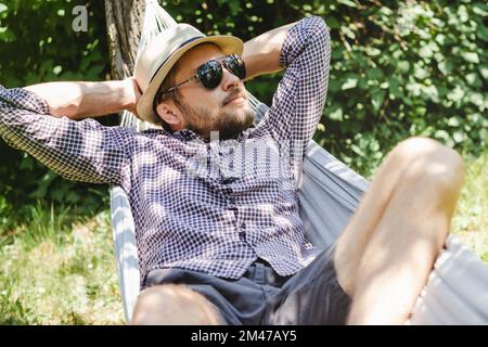 Bearded man in hat and sunglasses lying on hammock in green garden. Stock Photo