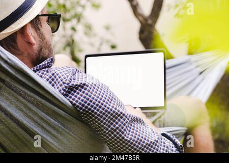 Businessman in hat and sunglasses lying down on hammock and using his laptop. Stock Photo