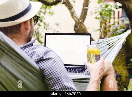 Laptop with blank screen and rear view man lying down in a hammock with a glass of water in his hand. Stock Photo