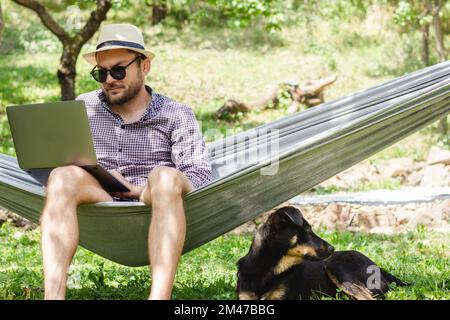 Man wearing hat lying down on hammock and using his laptop, dog lying near the owner on the ground. Stock Photo