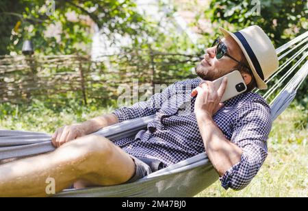 Bearded man in hat and sunglasses calling phone while lying on the hammock in the garden. Stock Photo