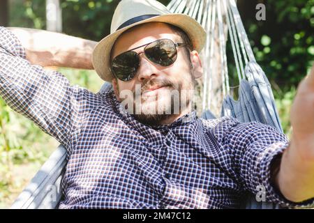 Man in straw hat and sunglasses lying down on hammock and taking a selfie. Stock Photo
