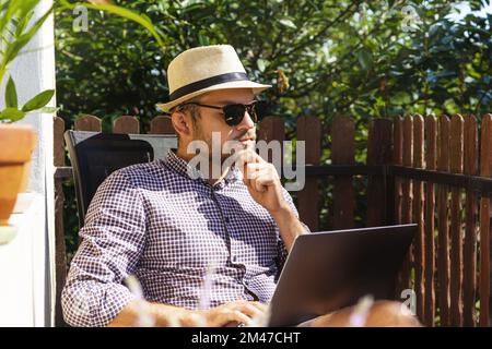 Adult bearded man wearing straw hat and sunglasses sitting on the balcony and using his laptop. Stock Photo