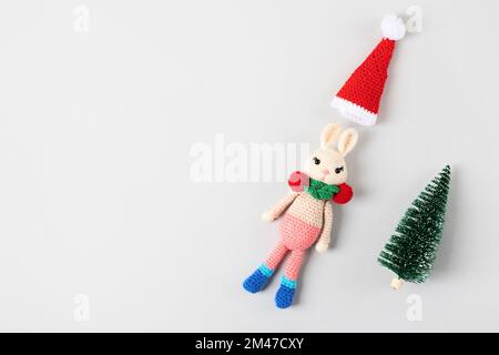 Knitted children's toy bunny with a Santa Claus hat and a small green Christmas tree on a gray background with a place for text, the concept of the ne Stock Photo