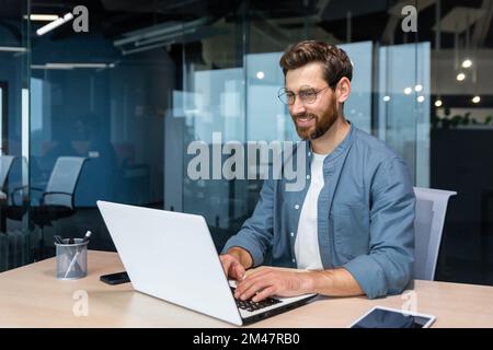 Smiling and cheerful businessman in casual shirt working inside modern office, mature programmer writing code using laptop. Stock Photo