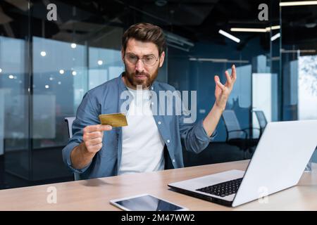 Cheated and disappointed businessman working inside office, man holding bank credit card and trying to make a purchase in online store, online fraud and deception. Stock Photo