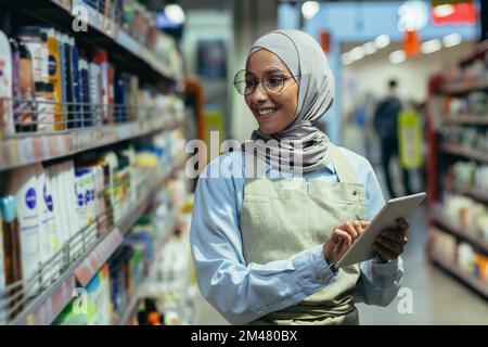 A woman checks products and goods in a supermarket, a Muslim woman in a hijab uses a tablet near a shelf with goods, a worker in an apron smiles at work. Stock Photo