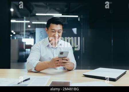 A young Asian businessman sits in the office at the table, holds the phone in his hands, types a message, reads the news, smiles. Stock Photo