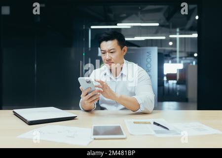 Worried young man Asian businessman sits in the office at the table, holds the phone in his hands, dials a message, is upset and reads bad news. Stock Photo