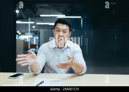 Online psychotherapy. Serious young Asian psychologist man sitting in the office at the desk and talking to the camera. Conducts a consultation with the patient remotely through a video call. Stock Photo