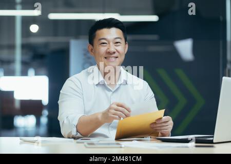 A young man, an Asian journalist, is sitting in the office at the table, holding an envelope with a letter, documents, an article in his hands. working on a laptop. He looks at the camera, smiles. Stock Photo