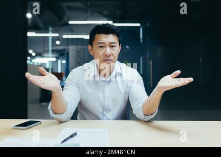 Worried and serious young Asian man, office worker sitting at the desk, spreading his hands, having problems, looking at the camera. Stock Photo