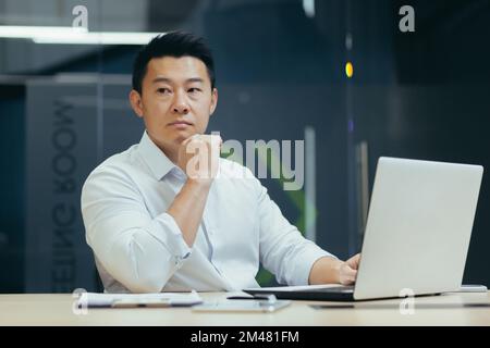Close-up photo. Portrait of a young serious handsome Asian lawyer, legal advocate. He sits thoughtfully in the office at a desk with a laptop, holds his head with his hand, looks to the side. Stock Photo