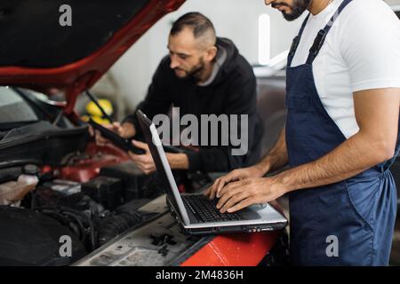 Cropped view of bearded men colleagues car technician mechanics repairing car problem of engine, during system checking detail, using laptop computer for maintenance and fixing in car garage. Stock Photo