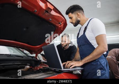 Young bearded men colleagues car technician mechanics repairing car problem of engine, during system checking detail, using laptop computer for maintenance and fixing in car garage. Stock Photo