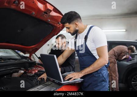 Young bearded men colleagues car technician mechanics repairing car problem of engine, during system checking detail, using laptop computer for maintenance and fixing in car garage. Stock Photo