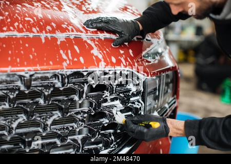 Close up photo of man hand in black protective gloves cleaning radiator grille in foam with special brush. Carwash and detailing. Washing machine at the station. Car washing concept Stock Photo