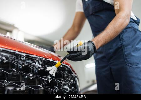 Closeup view of hand with special yellow brush of wash worker cleaning red crossover grille in a vehicle detailing workshop. Auto service industry. Stock Photo