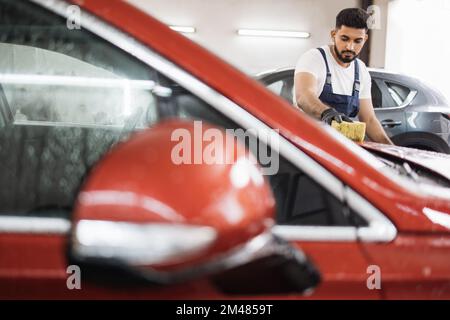 Car wash and clean with shampoo and sponge. Young concentrated bearded man worker, wearing blue overalls and black gloves washing soapy red car hood with yellow sponge on a car wash. Stock Photo