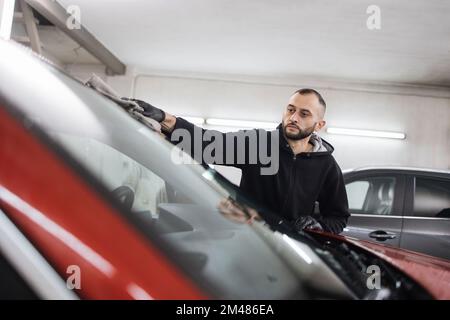 Car detailing and cleaning concept. Good-looking young male car wash worker, wearing black gloves, wipes and polishes car windshield window with microfiber cloth. Stock Photo