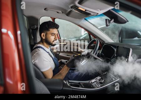 Auto cleaning service and detailing concept. Handsome young bearded man in blue uniform overalls cleaning interior of the car with hot steam cleaner. Stock Photo
