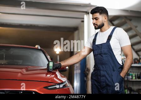 Young attractive man, professional service specialist testing thickness of the car paint in protective gloves . Car paint coating metal parts testing before buying. Used vehicle purchase risks. Stock Photo