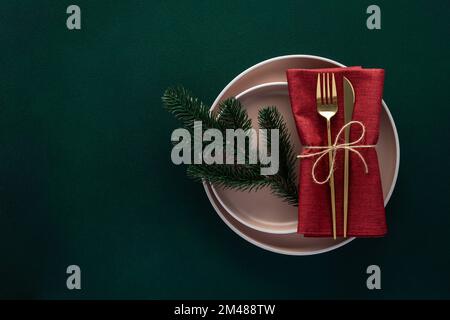 Christmas or New Year dinner table setting with empty plate and golden cutlery on dark green background, card or menu template with fir tree. Copy spa Stock Photo