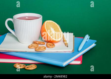 Notebooks, a cup of tea and an orange on a green background, copy space. Stock Photo