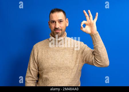 Hispanic man wearing brown turtleneck unshaven happy smiling and showing ok sign at camera isolated over blue background Stock Photo