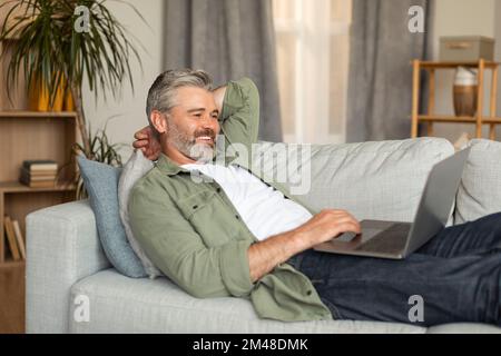 Smiling elderly caucasian man typing on computer, resting, lying on sofa and watching video in living room Stock Photo