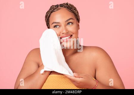 Attractive african american body positive woman drying face with soft towel, caring for skin over pink background Stock Photo