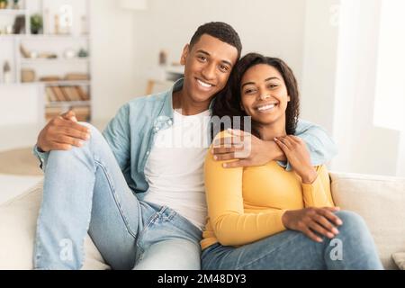 Cheerful handsome young african american man hugging his wife, resting on sofa, enjoy weekend Stock Photo