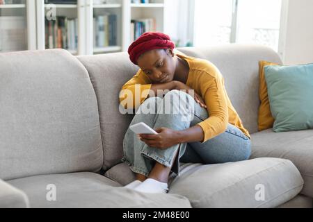 Stressed black woman sitting on sofa with cellphone Stock Photo