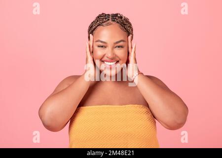 Portrait of excited black body positive woman touching face and enjoying her smooth skin, pink studio background Stock Photo