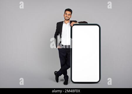 Business Ad. Smiling Young Arab Businessman Leaning At Big Blank Smartphone Stock Photo