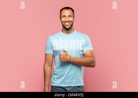 Happy african american man showing thumb up and cheerfully smiling at camera, posing isolated on pink studio background Stock Photo