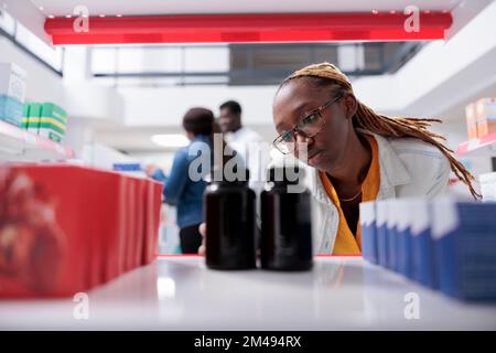 African american woman taking medication bottles from drugstore shelf, selective focus. Pharmacy client and seller talking in background, pharmacist putting pills packages close up Stock Photo