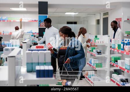 African american buyer choosing heart medication in drugstore, reading instruction, standing in pharmacy aisle. Woman customer buying vitamins, pharmacist in background, all black team Stock Photo