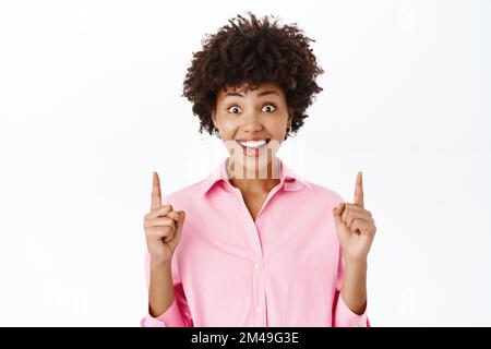 Enthusiastic african american girl points finger up, shows advertisement on top. Businesswoman in pink shirt shows promo text Stock Photo