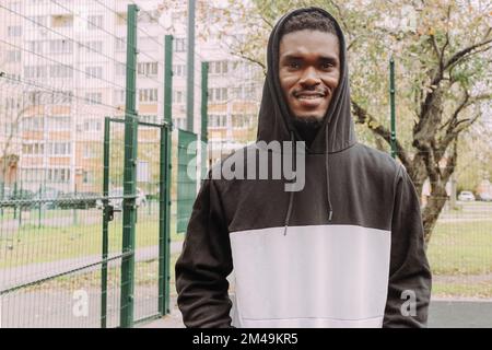 Smiling young black man in two-tone hoodie over sport court backdrop. Close up portrait of smiling African guy looking at camera Stock Photo