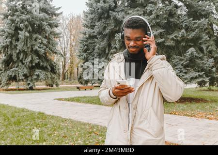 Handsome black man in headphones searching songs in playlist over park background. Smiling guy in warm casual clothes with wireless earphones Stock Photo