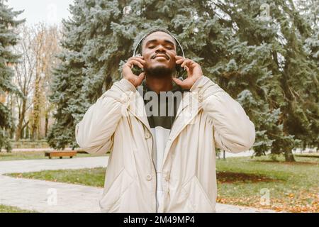 Portrait of handsome black man in wireless headphones over park backdrop. Smiling African guy with closed eyes in warm clothes and in white earphones Stock Photo
