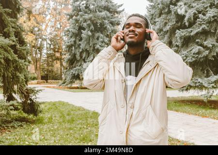 Handsome black man in wireless headphones listening to music outdoors. Smiling guy with closed eyes holding his hands on white earphones Stock Photo