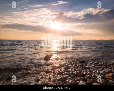 Autumn sunset view across a stretch of water with waves beating boulders on the bank under the blue sky, sun beaming between pink clouds creating a light path Stock Photo