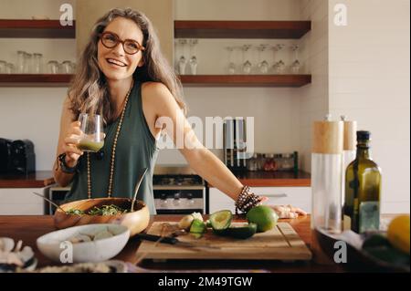 Healthy senior woman smiling while holding some green juice in her kitchen. Mature woman serving herself wholesome vegan food at home. Happy woman tak Stock Photo