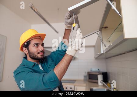 Focused professional worker in uniform is assembling furniture on kitchen. Repair concept Stock Photo