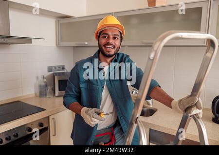 Smiling handyman is standing on ladder in kitchen and holding a screwdriver Stock Photo