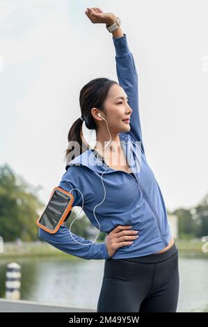 Portrait, Active and charming millennial Asian female in active sportswear listening to music through her earphones while stretching her arms. Stock Photo
