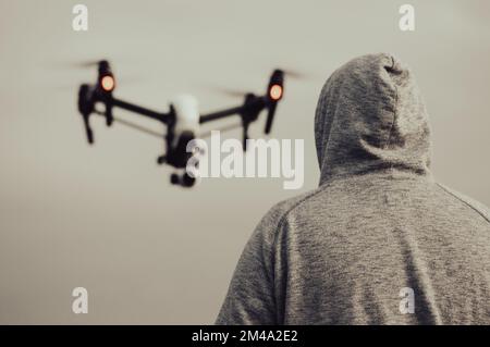 A back shot of a hoodie male looking at a flying blur drone in the gray sky Stock Photo
