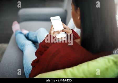 Thinking about what to post online. Rearview shot of an unrecognizable woman using a cellphone while chilling on the sofa in the living room at home. Stock Photo